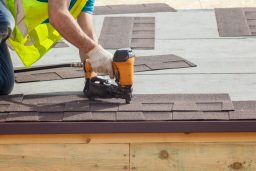 Clive roofing worker putting the asphalt roofing (shingles) with nail gun on a new frame house