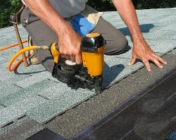 A new frame house's asphalt roofing (shingles) are being installed by an Ankeny roofer using a nail gun.